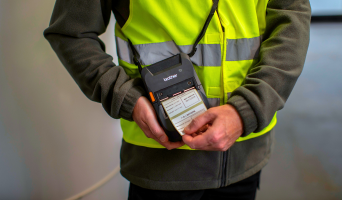 A person in a yellow safety vest prints labels from a handheld label printer while performing Field Service tasks.
