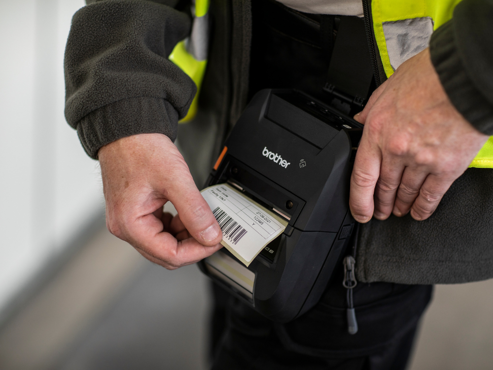 A person in a high-visibility jacket uses a portable Brother label printer to print a barcode label during Field Service tasks.