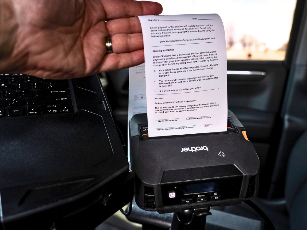 A hand holds a printed document from a Brother printer inside a vehicle next to a laptop, illustrating mobile printing solutions ideal for Public Sector professionals.