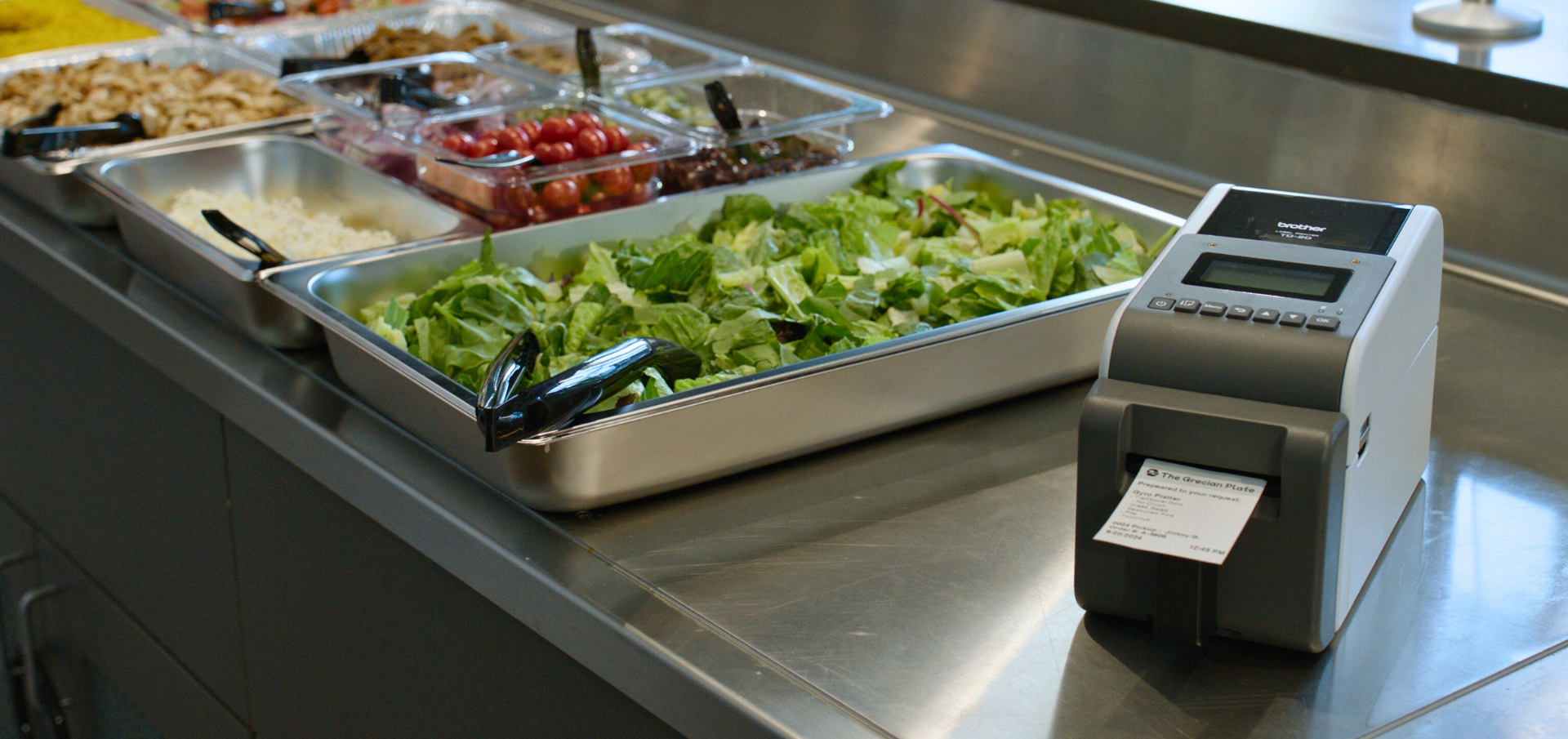 A salad bar with fresh greens and toppings next to a label printer on a stainless steel countertop.
