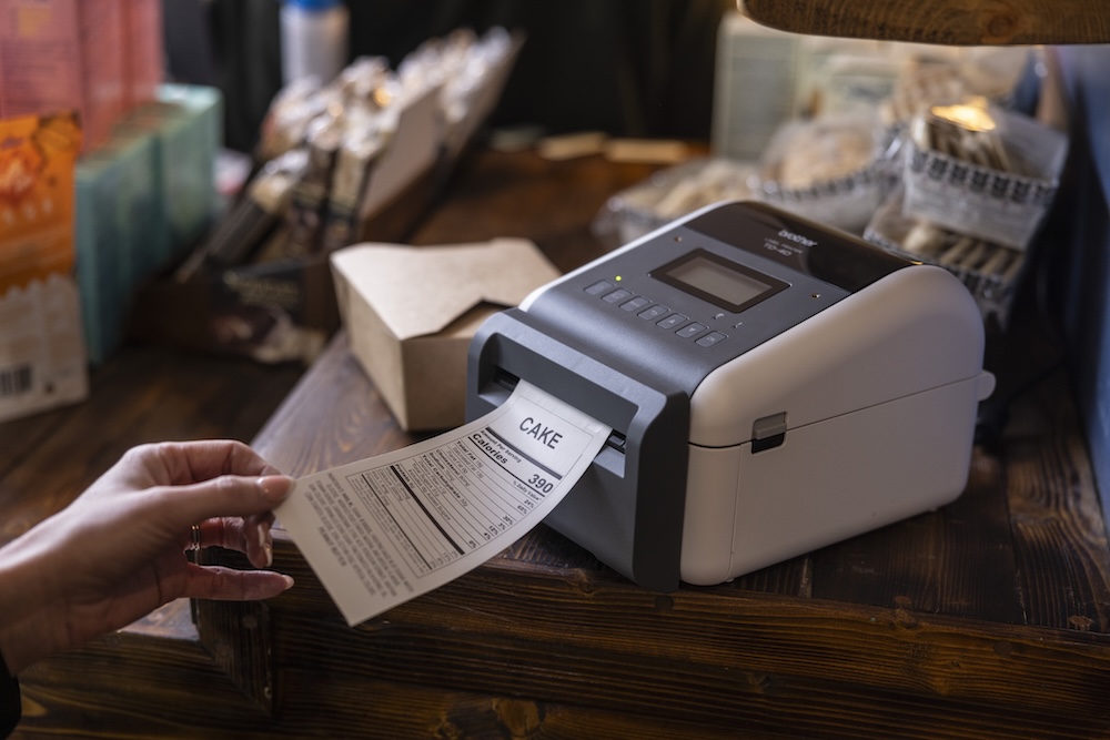 A hand pulls a printed receipt labeled CAKE from a receipt printer on a wooden counter.