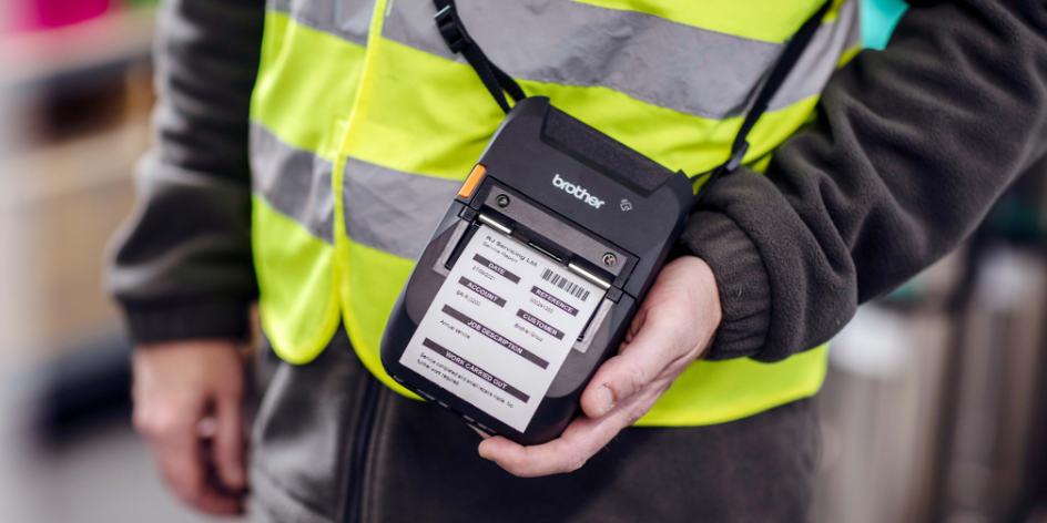 A person in a yellow safety vest holds a portable Brother label printer—ideal among printers for vehicles—displaying freshly printed labels.