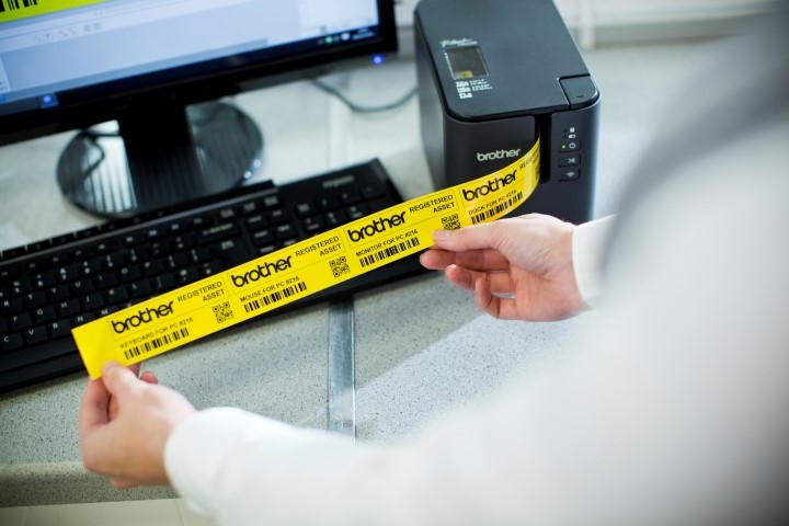 A person holds a printed yellow label in front of a keyboard and Brother label printer on a desk, showcasing industrial labeling efficiency.