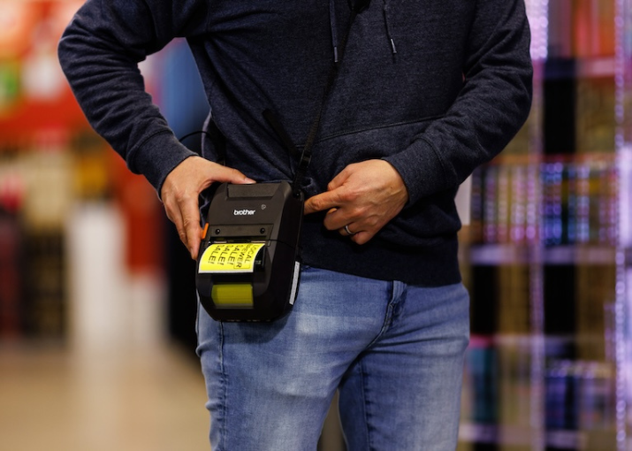 Man in refrigerated convenience store isle wearing a Brother RuggedJet 3 inch printer on his hip, printing a sale label.