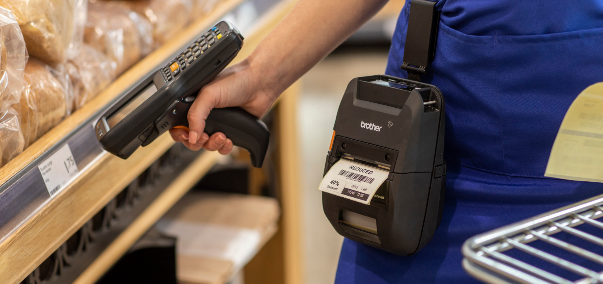 Person in blue apron scanning a label with a handheld barcode scanner near Price Label Printers in a grocery store bakery aisle.