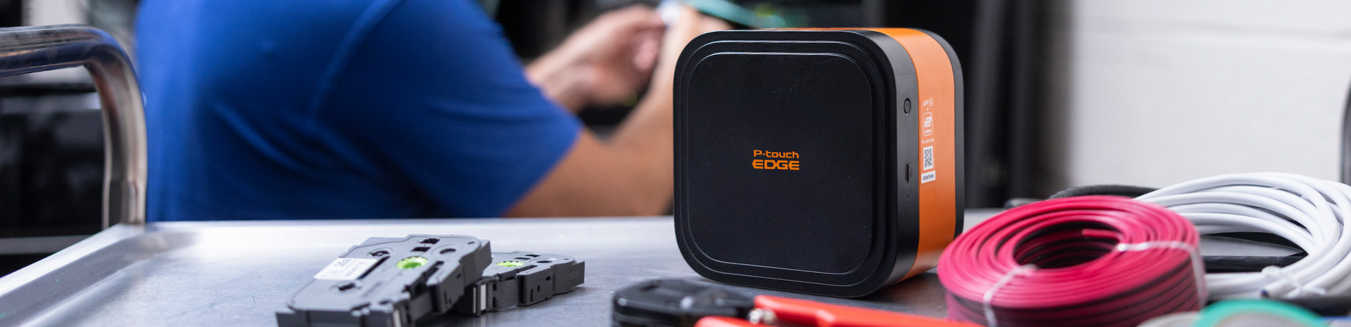 A P-touch EDGE 720BT industrial labeling printer rests on a table among cables and tools, with a person blurred in the background.