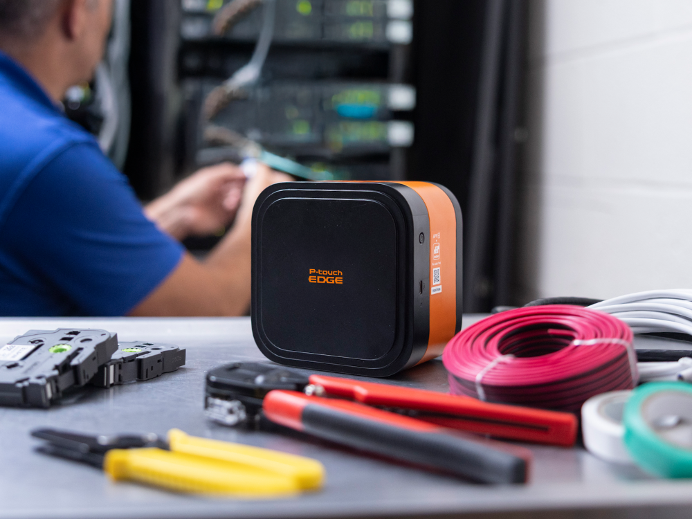 A technician works in a server room; network tools, cables, and a P-touch EDGE 720BT label printer are on a table in the foreground.