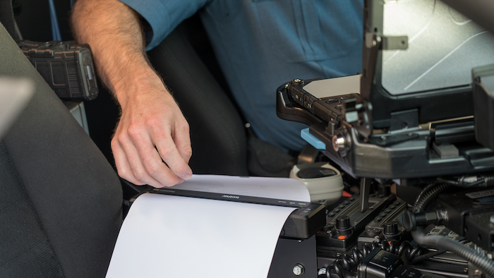 A person’s hand uses a portable printer inside a vehicle, next to a mounted computer and various equipment