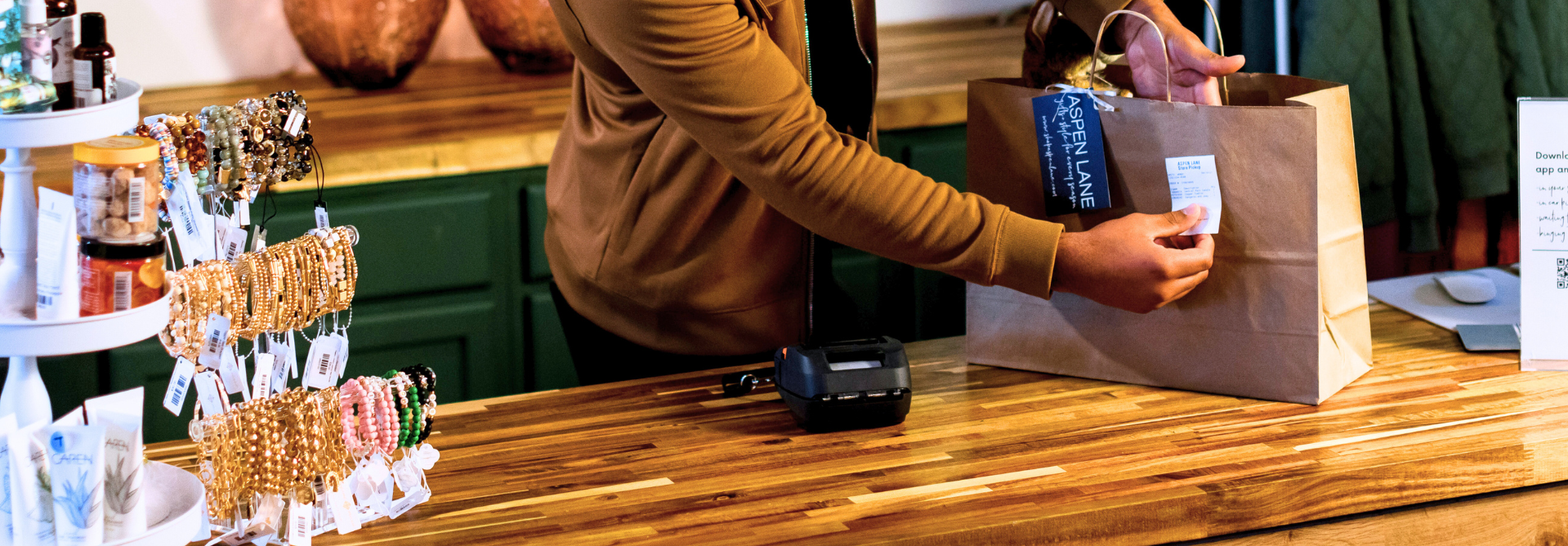 Someone uses the RuggedJet 3255WB-LF mobile printer to label a paper shopping bag on a wooden counter next to jewelry and product displays.