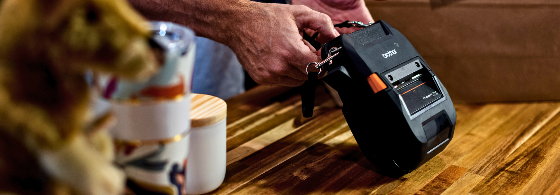 A person holds the RuggedJet 3255WB-L label printer on a wooden table, surrounded by mugs and a plush toy.