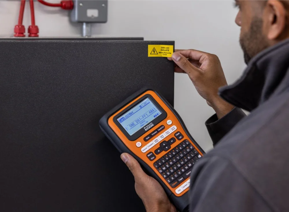 A person is holding an orange and black label maker and applying a yellow warning label that reads Risk of electric shock on a dark gray box. Red cables are visible in the background.