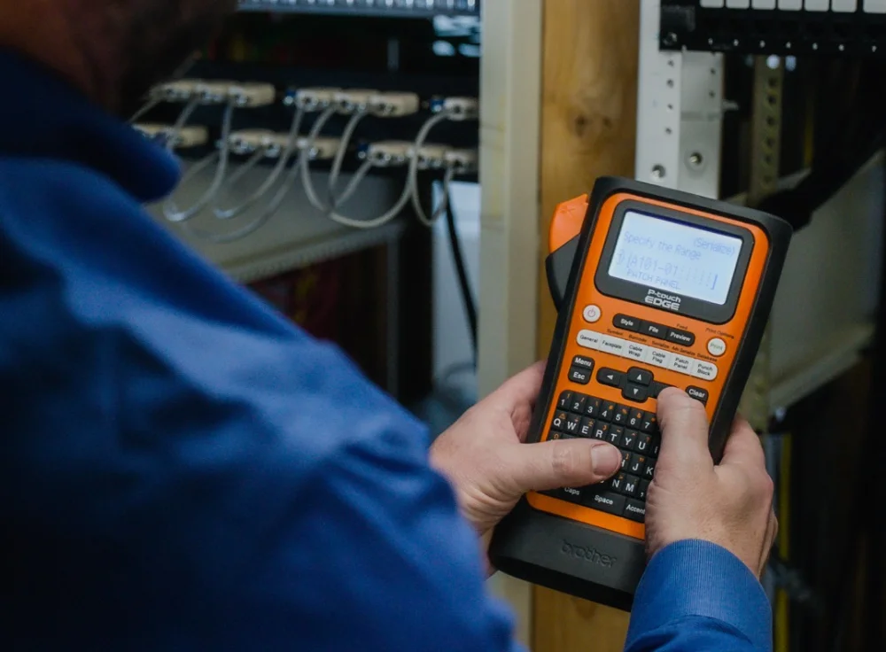 A person in a blue shirt uses an orange label printer to create labels for heat shrink tubing. They are standing in front of a rack filled with cables and electronic equipment.