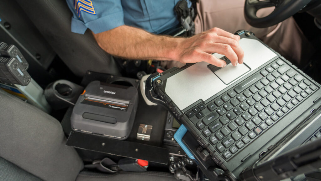 A police officer uses a RuggedJet 4200 rugged laptop in a patrol car, with a printer and radio visible nearby—demonstrating smarter upgrades for the modern mobile workforce.