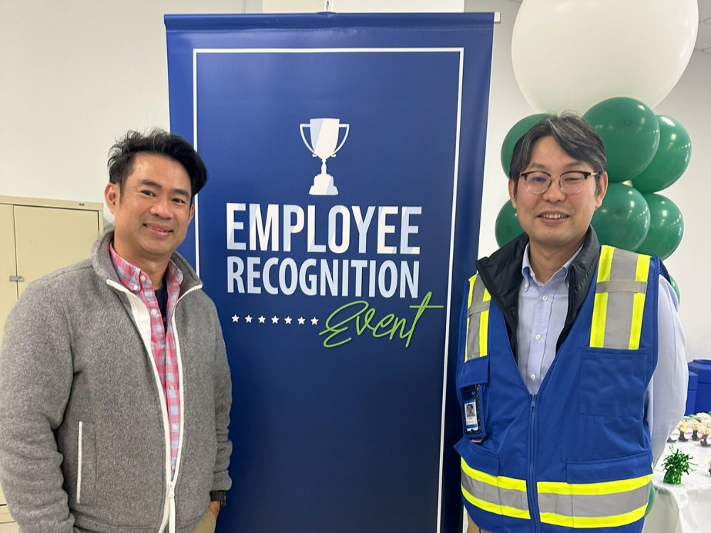 Two men stand smiling in front of an Employee Recognition Event banner with balloons in the background, celebrating recent management changes at Brother International Corporation.