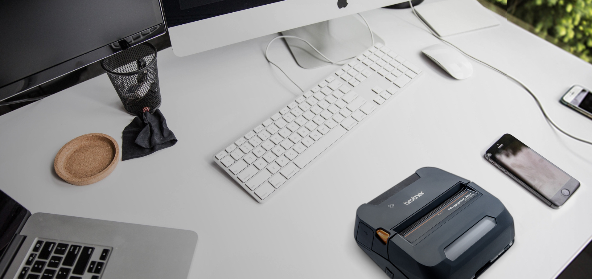 A tidy white desk with computer monitors, keyboard, mouse, phone, a Brother label printer, and tools ready for Mobile Deploy.