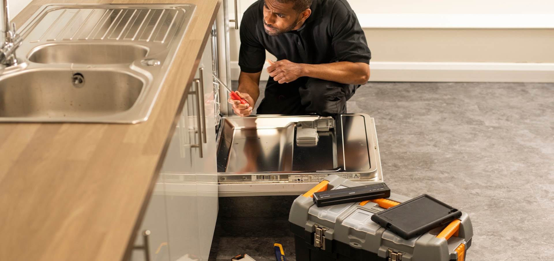 A man kneels to repair a dishwasher in a kitchen, with tools and a toolbox on the floor nearby.