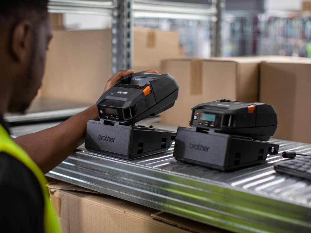 A person placing a portable Brother printer onto a charging dock in a warehouse setting.