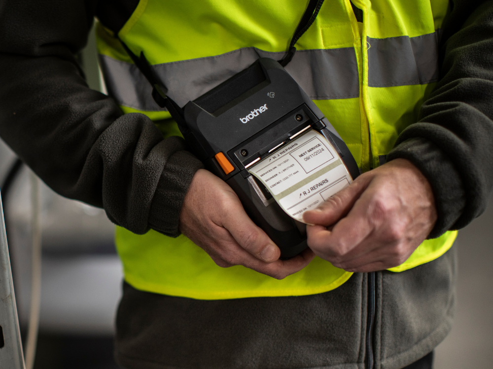 Person in a yellow safety vest prints a label using a handheld Brother label printer.