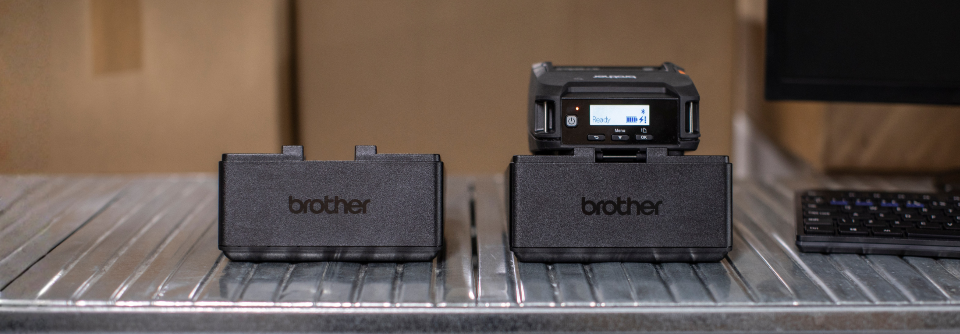 Two Brother label printer devices on a metal surface, one with a display screen, next to a computer keyboard.