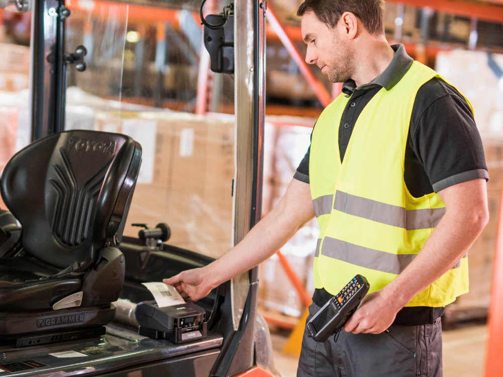 A man in a yellow safety vest scans a label inside a warehouse near a forklift.