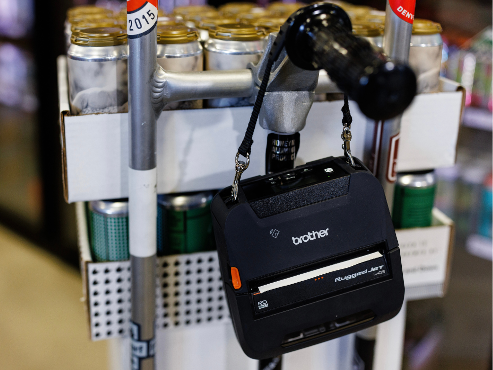 A Brother RuggedJet printer hangs on a cart handle in front of a pack of canned drinks in a store.