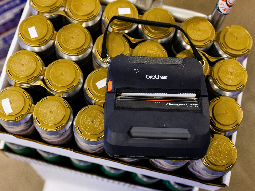 A Brother RuggedJet printer sits on top of a case of cans with gold lids in a cardboard tray.