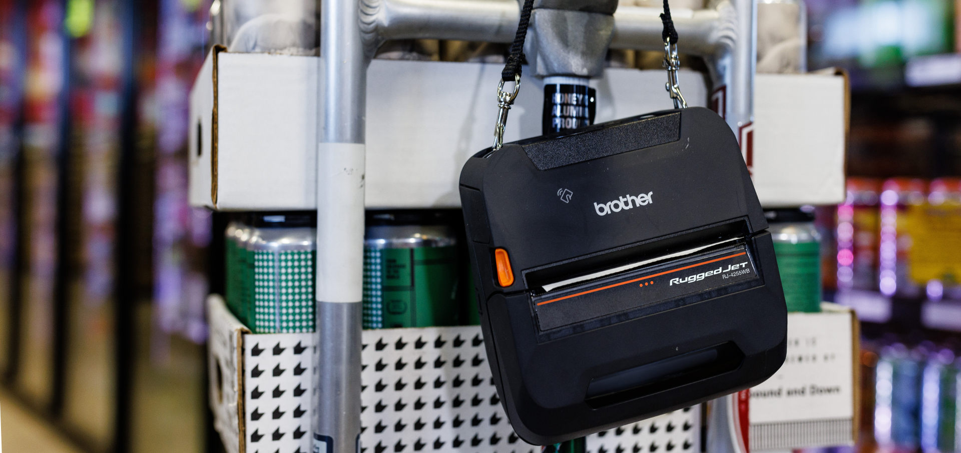 A Brother RuggedJet printer hangs on a cart loaded with canned drinks in a store aisle.
