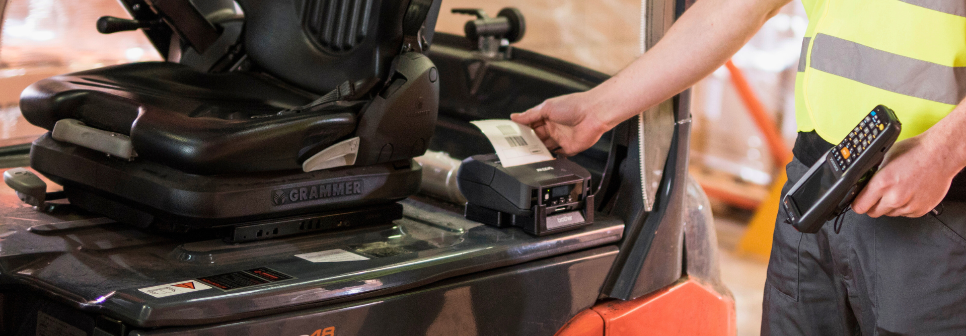 A worker in a safety vest collects a printed label from a machine on a forklift, holding a handheld scanner.
