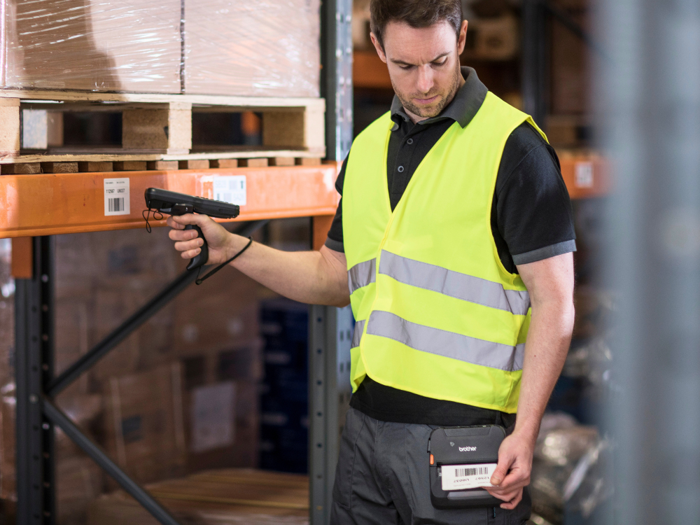 A warehouse worker in a yellow vest scans a barcode on a shelf using a handheld scanner.
