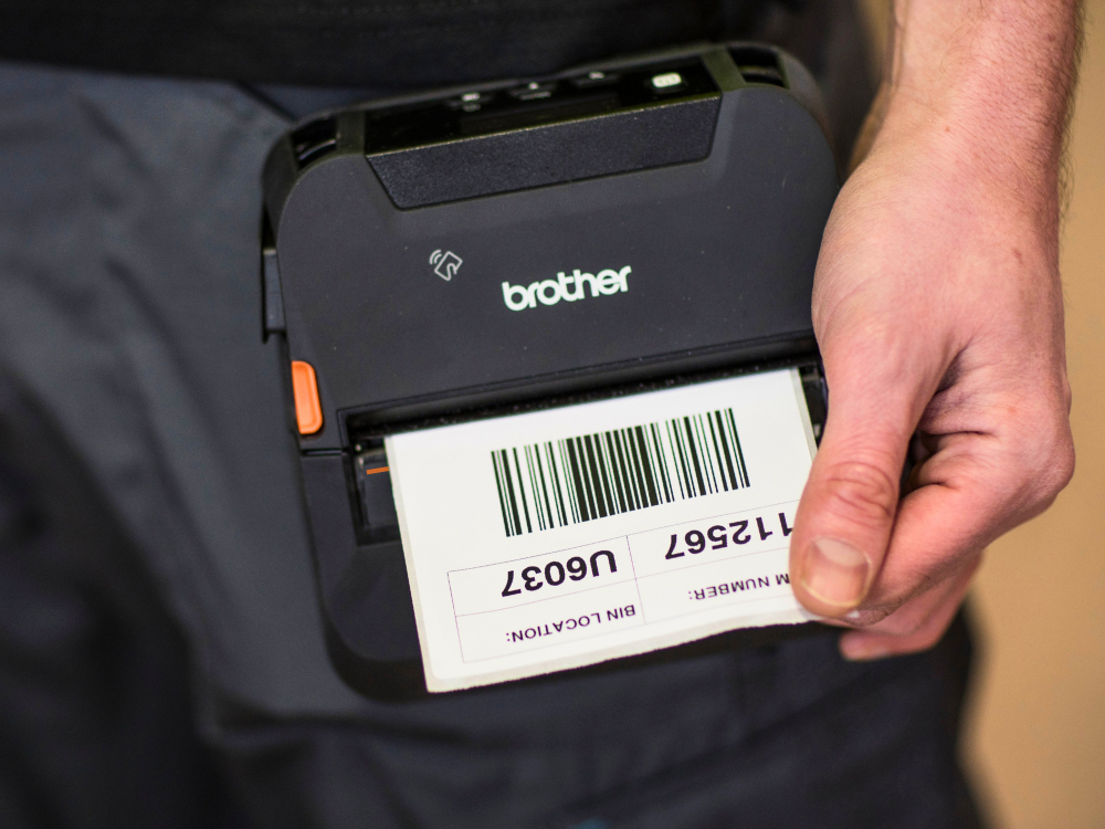 A person holds a Brother label printer printing a barcode label with numbers and text.