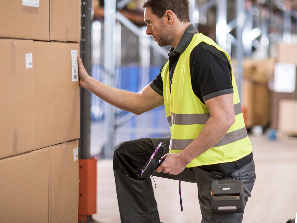 A man in a yellow safety vest uses the RuggedJet 4235B-LF mobile printer while scanning barcodes on cardboard boxes in a warehouse.