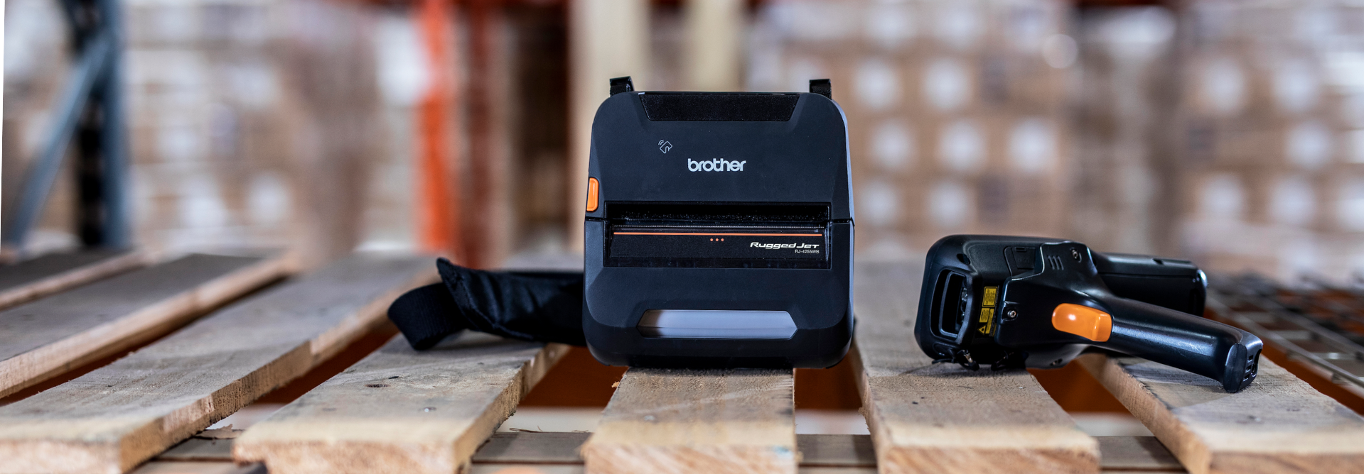 A Brother label printer and two barcode scanners resting on a wooden pallet in a warehouse setting.