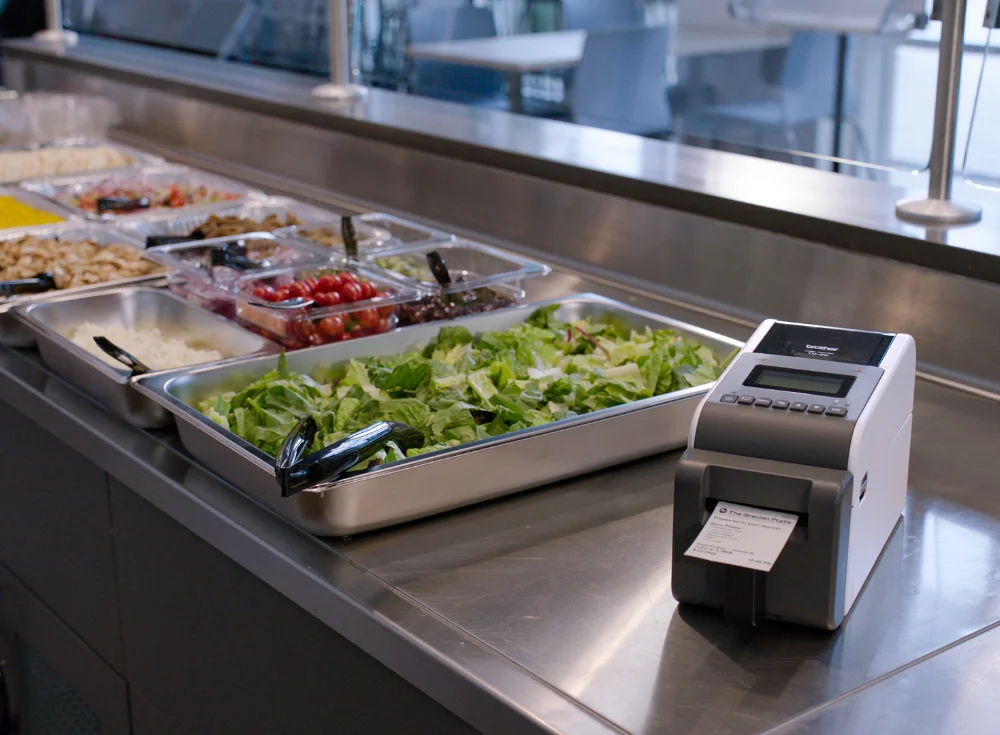 Self-serve salad bar with various ingredients and a label printer on a metal counter.