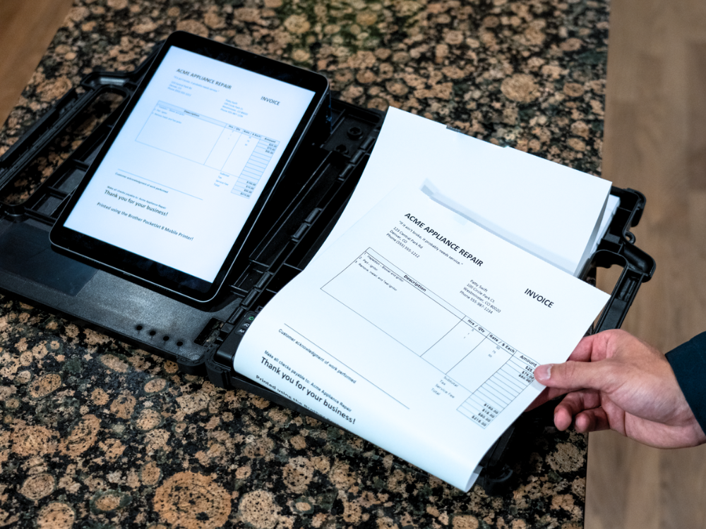 Person holding a printed invoice—produced using thermal printing—next to a tablet displaying the same invoice on a speckled countertop.