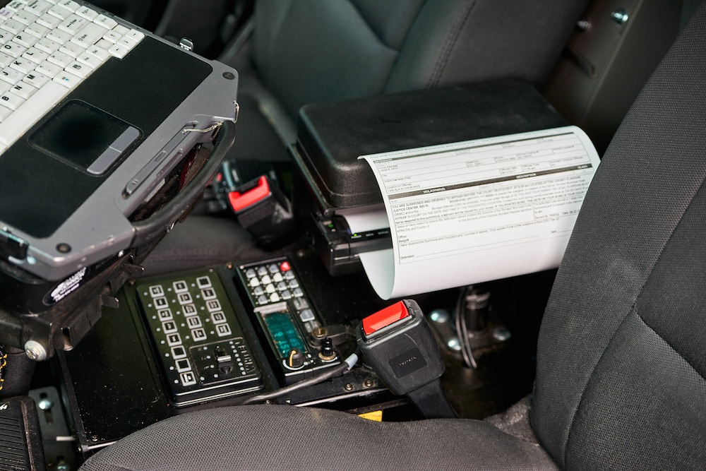 A police car interior with a mounted laptop, printer with paper utilizing Understanding Direct Thermal Printing Technology, radio, and control panel between seats.