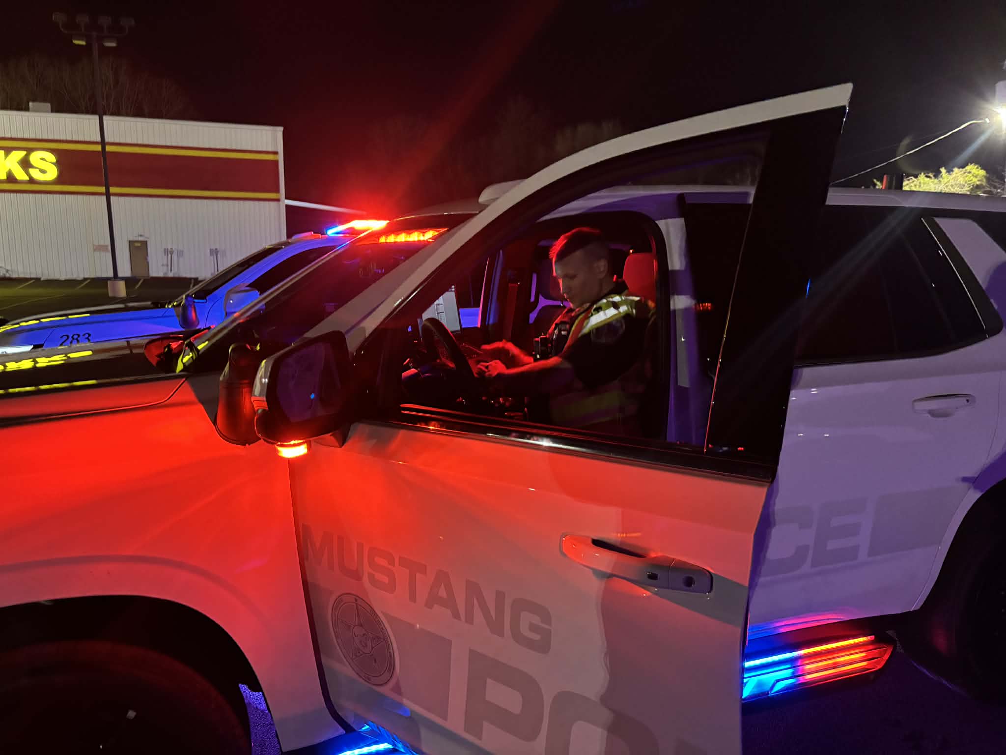 Police officer sits inside a Mustang Police vehicle at night with emergency lights flashing.